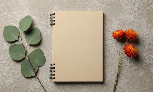 Cream colored spiral notebook placed on textured wall with green leaves and orange flowers for aesthetic display