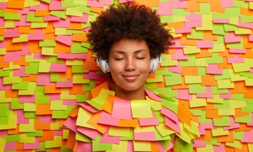 Relaxed ordinary ethnic woman listens pleasant audio track in headphones, keeps eyes shut, enjoys every sound, popular music in playlist, sticks out head through wall with colorful stickers.