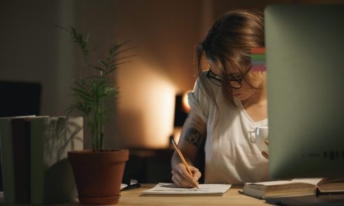 Image of concentrated young woman designer sitting indoors at night writing notes using computer. Looking aside.