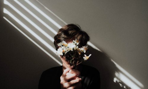 A male holding a small bouquet of flowers in front of his face with light lines shining on him near a white wall