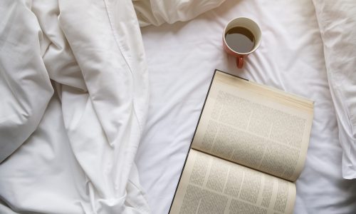 Topview point of red cup of coffee, open book, white blanket and white pillow on white bed. Sunlight effect.