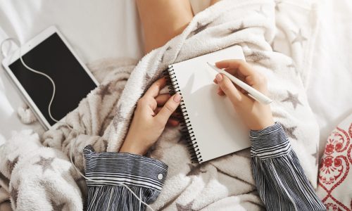 There is no place like bed. Creative and relaxed woman lying under blanket, listening music in earphones via digital tablet, making notes while working from home. Girl uses imagination.
