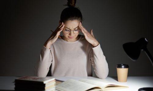 Tired stressful woman keeps hands on head, suffers from headache after working all night, tries to conecntrate, continue reading, dressed in casual clothes, wears transparent glasses enjoys cappuccino