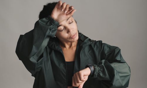 Young wet African American woman in wireless earphones checking her run result on smartwatch looking tired over gray background. Modern technology